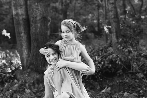 Czarno-białe zdjęcie uśmiechniętej kobiety niosącej na barana małą dziewczynkę w sukience, na tle leśnego krajobrazu.