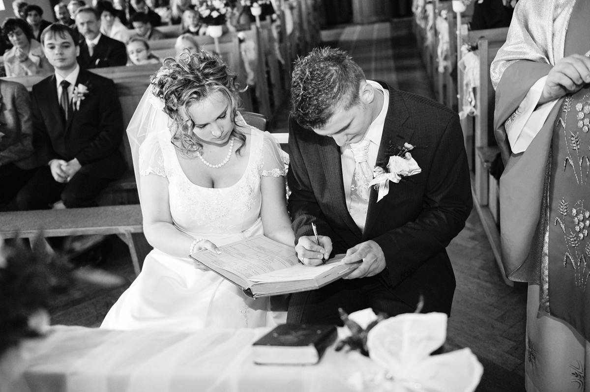 Czarno-białe ujęcie pary młodej podpisującej akt małżeństwa w kościele, panna młoda w koronkowej sukni z perłami, pan młody w ciemnym garniturze z butonierką, w tle widoczni goście.