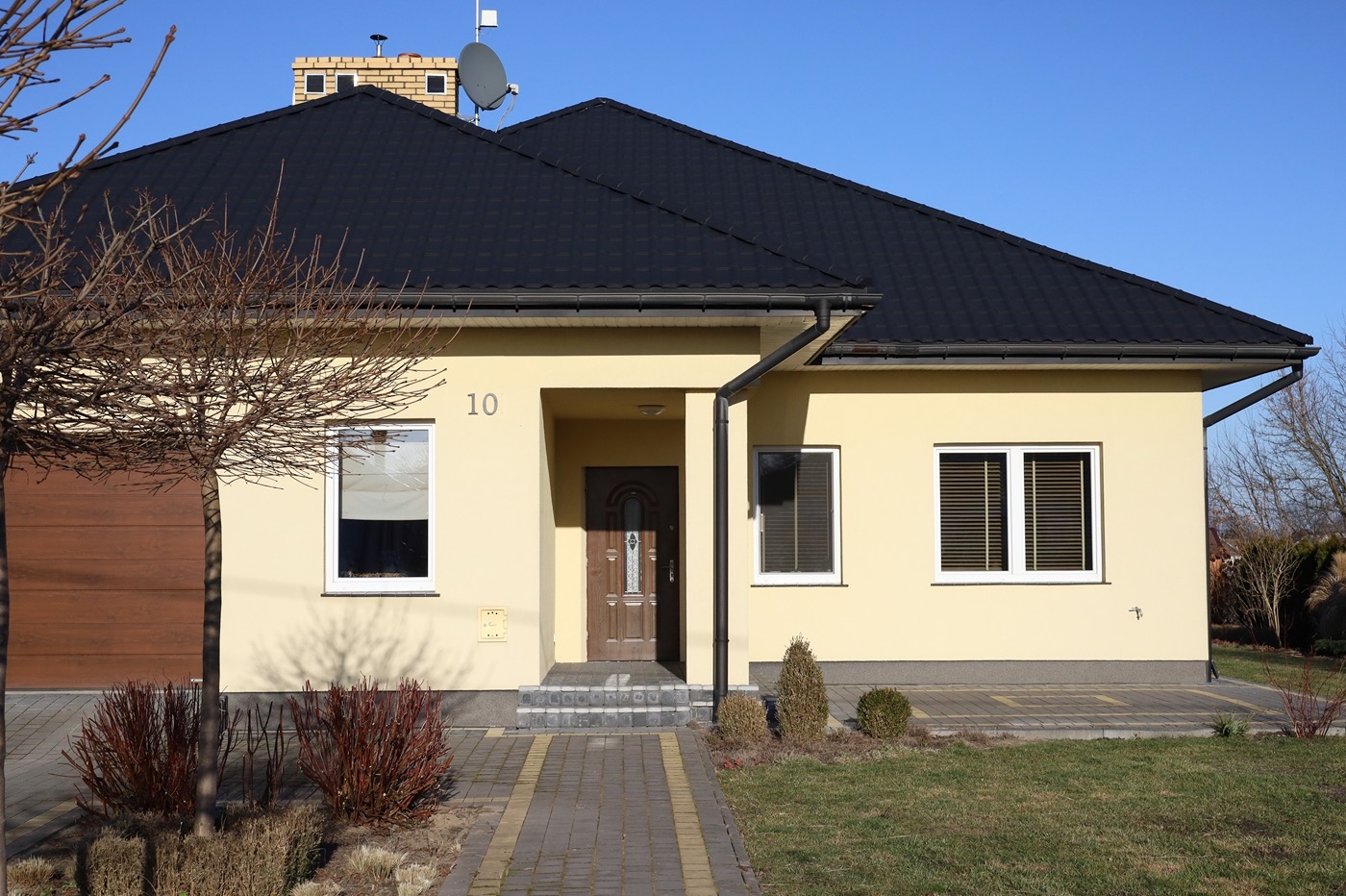 Elewacja domu jednorodzinnego z czarnym dachem i numerem 10 nad wejściem, widziana z poziomu chodnika.