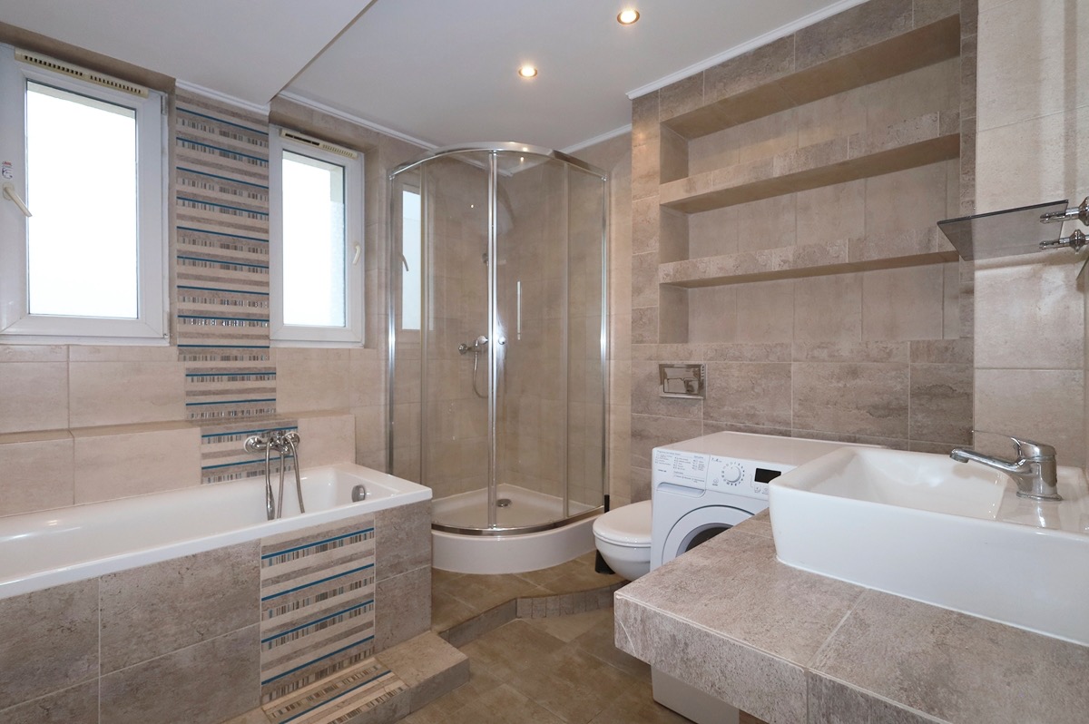 Jasna łazienka z wanną, kabiną prysznicową, umywalką nablatową i pralką, wykończona beżowymi płytkami z poziomymi paskami.