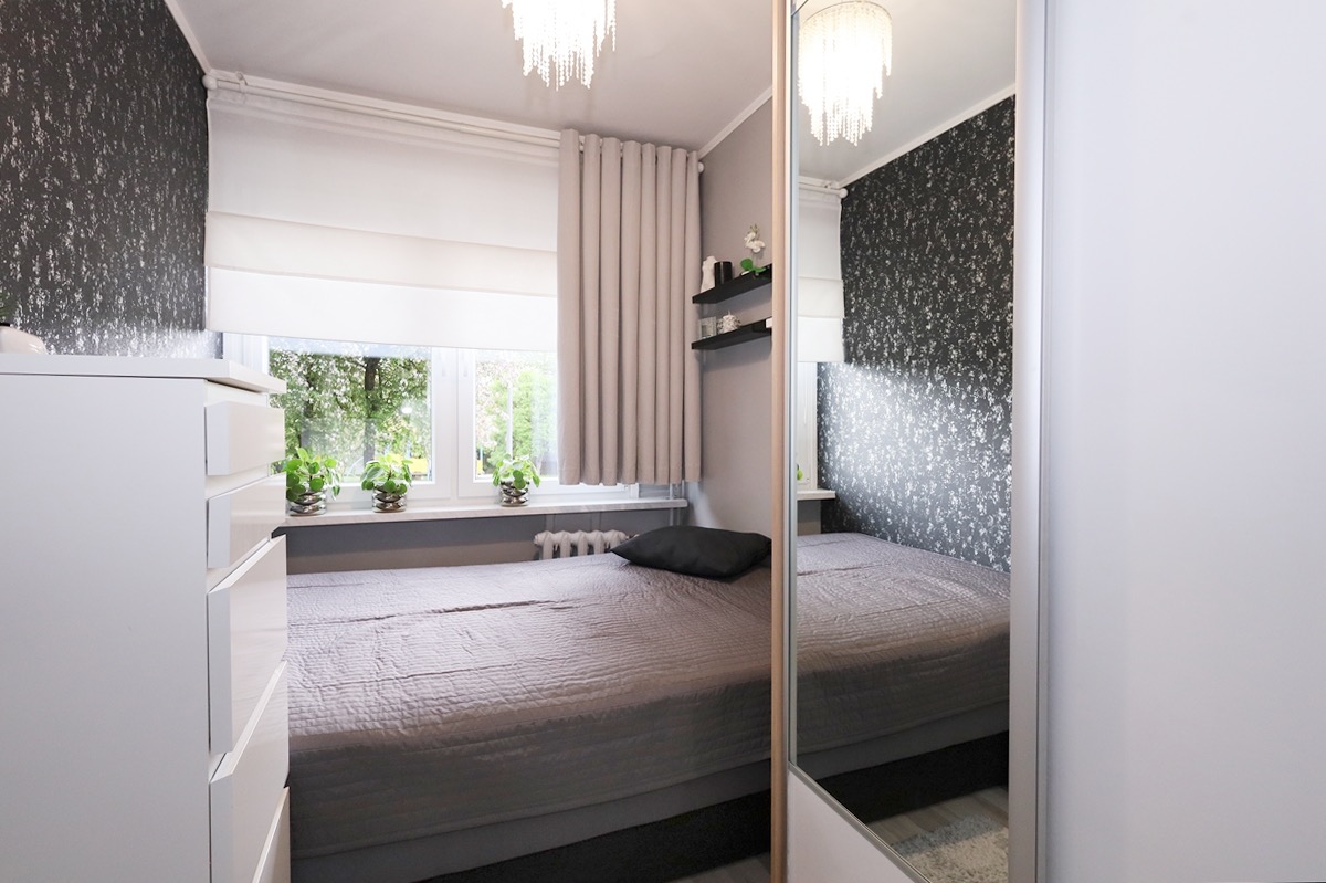 Nowoczesna sypialnia z szarą pościelą, wzorzystą tapetą, białymi meblami i lustrzanym frontem szafy przesuwnej, oświetlona lampą kryształową