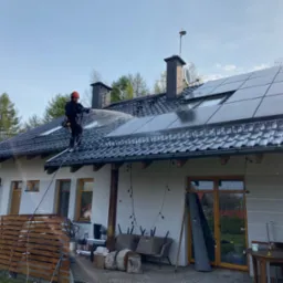 Pracownik w uprzęży zabezpieczającej spryskuje wodą pod ciśnieniem dach pokryty panelami słonecznymi i dachówką, w tle widoczne drzewa i budynek z oknami.