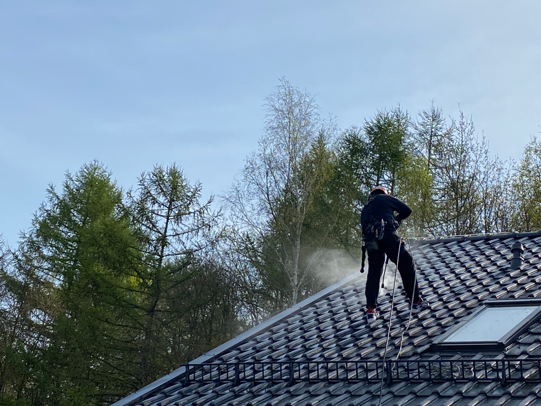 Pracownik w uprzęży, stojący na ciemnoszarym dachu z dachówką, spryskuje go wodą pod ciśnieniem; w tle drzewa i błękitne niebo.