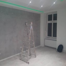Łukbud - Wnętrze pokoju po nałożeniu gładzi na ścianę, z widoczną aluminiową drabiną, oświetleniem LED w suficie i grzejnikiem pod oknem.