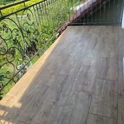 Łukbud - Wyłożony drewnopodobnymi płytkami balkon z metalową, ozdobną balustradą na tle zieleni.