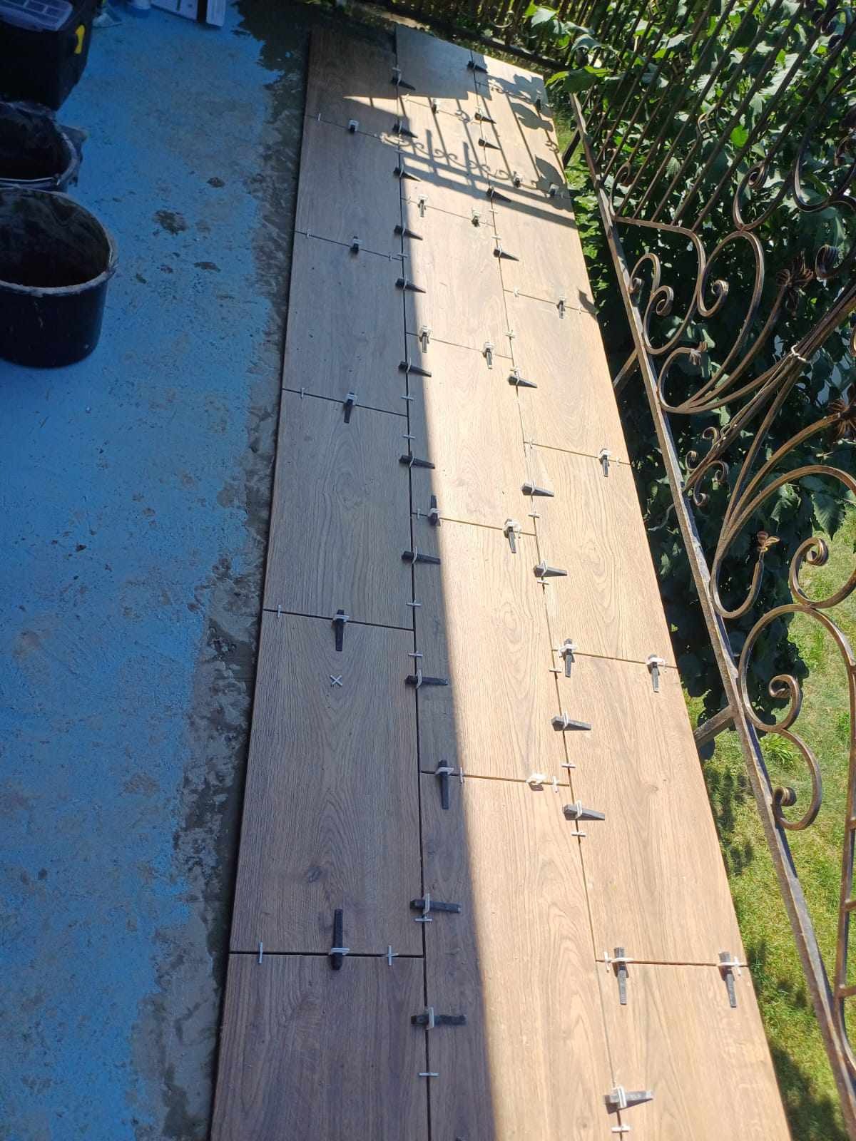 Układanie płytek imitujących drewno na zewnątrz, z użyciem krzyżyków dystansowych, widoczna balustrada z metaloplastyki i wiadro z zaprawą.