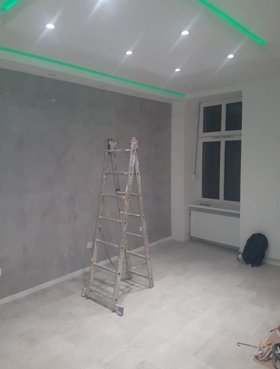 Wnętrze pokoju po nałożeniu gładzi na ścianę, z widoczną aluminiową drabiną, oświetleniem LED w suficie i grzejnikiem pod oknem.