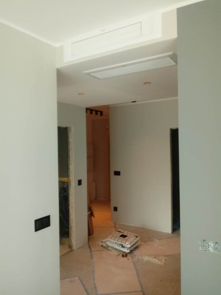 Wnętrze w trakcie prac wykończeniowych: szare ściany, wystające kable elektryczne, zabezpieczona podłoga, widoczny klimatyzator pod sufitem i opakowanie po płytkach.