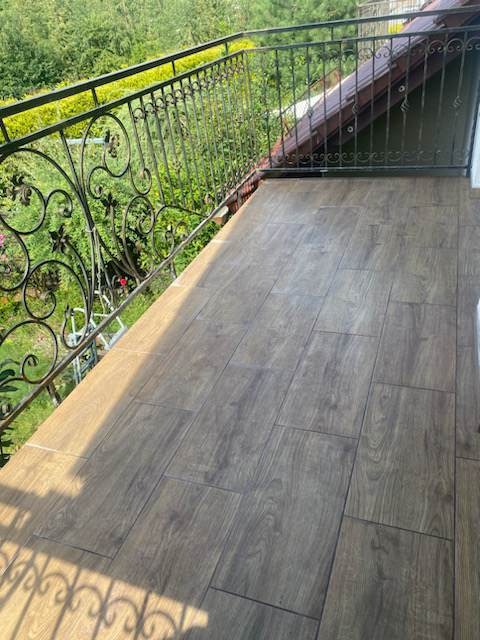 Wyłożony drewnopodobnymi płytkami balkon z metalową, ozdobną balustradą na tle zieleni.