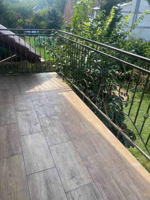 Wyłożony płytkami imitującymi drewno balkon z metalową, ozdobną balustradą, widok na zieleń ogrodu.