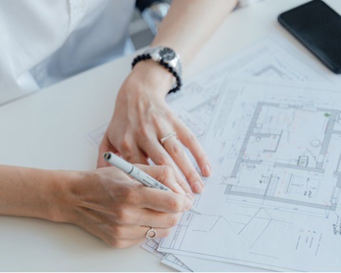 Kobieta w białej koszuli nanosi poprawki na plan architektoniczny domu, używając szarego markera. Na nadgarstku widoczny zegarek z czarnym paskiem z koralików. W tle leży czarny telefon.