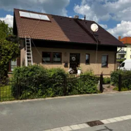 Dom jednorodzinny z częściowo pomalowanym na brązowo dachem, zabezpieczonym plandeką fragmentem, drabina oparta o dach, widoczna antena satelitarna i przewody.