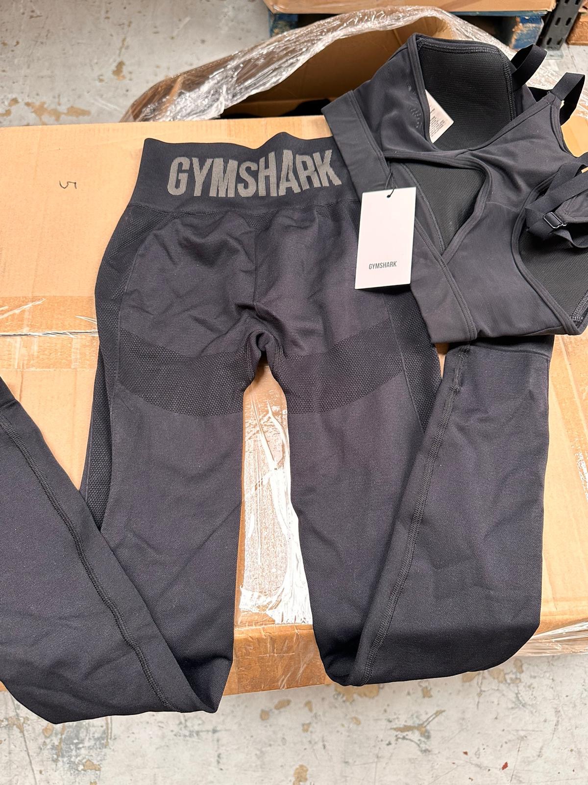Czarne legginsy i sportowy biustonosz marki Gymshark z metką, leżące na kartonowym podłożu.