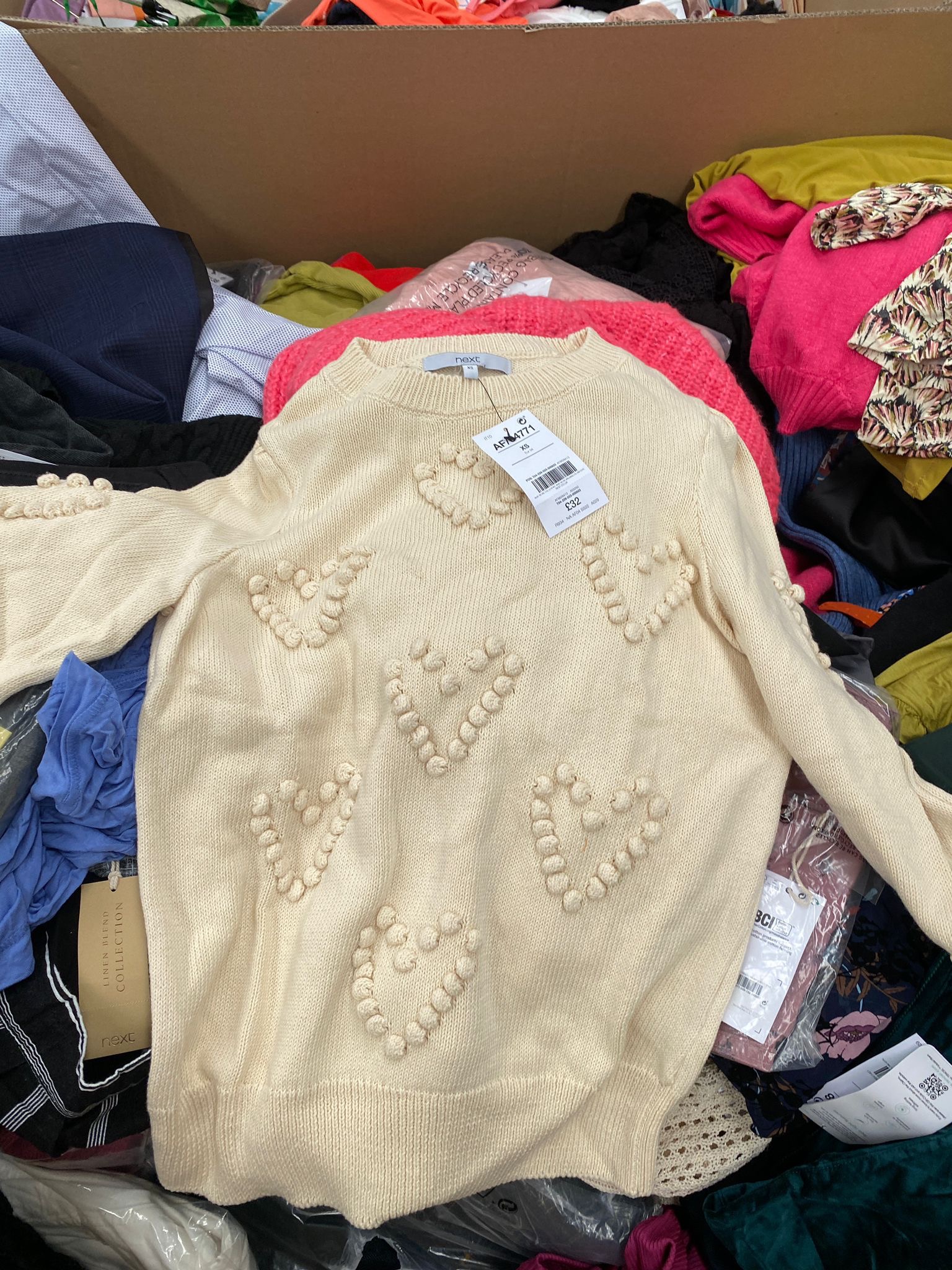 Kremowy sweter damski z wypukłymi sercami, widoczny w kartonie z innymi ubraniami, z metką cenową i informacjami o rozmiarze XS.