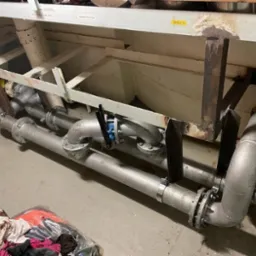 Metalowe rury przemysłowe z zaworem pod sufitem, zamontowane w hali, widoczne ślady spawania i mocowania konstrukcji wsporczej, ubrania robocze na podłodze.