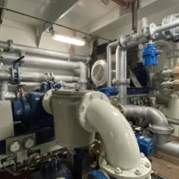 Złożona instalacja przemysłowa z rurami, zaworami, pompami i manometrami wewnątrz budynku, oświetlona lampą jarzeniową.