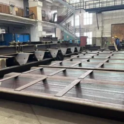 Wnętrze hali produkcyjnej z ułożonymi elementami konstrukcji stalowych w trakcie obróbki; widoczne iskry podczas szlifowania metalu, a w tle pracownicy w kaskach.