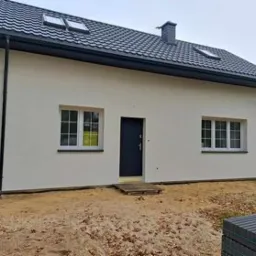 Nowoczesny dom jednorodzinny z białymi oknami PCV i ciemnymi drzwiami w Mińsku Mazowieckim, widok z zewnątrz na elewację i teren wokół domu w trakcie prac wykończeniowych.