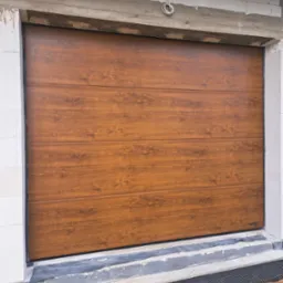 Zamknięta brama garażowa segmentowa z paneli w odcieniach brązu imitujących drewno, widoczna od strony zewnętrznej, osadzona w surowym, niewykończonym murze z betonu komórkowego.