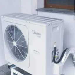 Biała zewnętrzna jednostka pompy ciepła marki Midea zamontowana na metalowym stelażu przy ścianie budynku z widoczną kratką wentylacyjną i rurami.