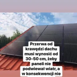 Panele słoneczne na czerwonym dachu z widocznym niebem i drzewami w tle, z nałożonym tekstem o konieczności zachowania odstępu od krawędzi dachu.