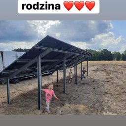 Nasz Prąd S.A. - Uśmiechnięta dziewczynka stoi obok metalowych słupków konstrukcji pod panele słoneczne na tle dwóch bawiących się dzieci i częściowo widocznego białego samochodu dostawczego.