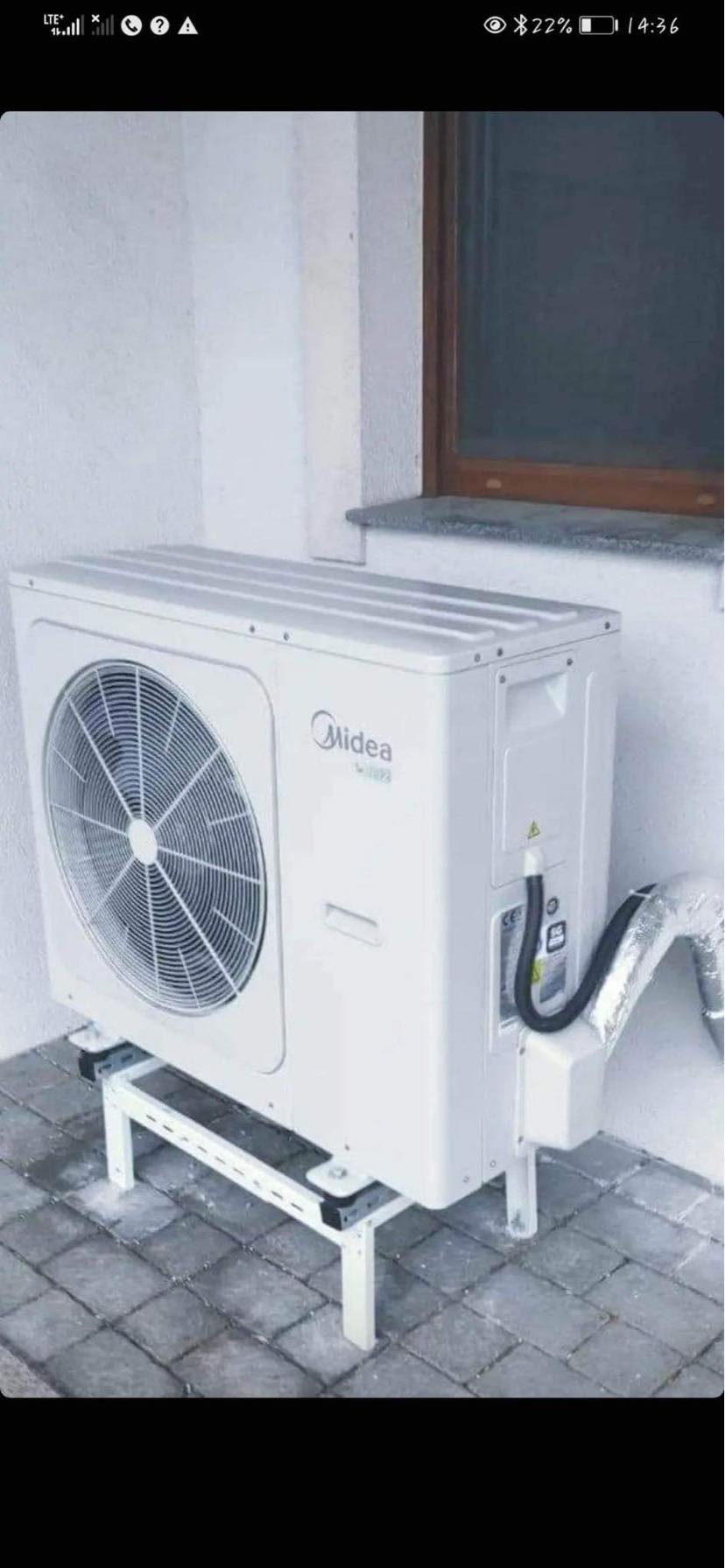 Biała zewnętrzna jednostka pompy ciepła marki Midea zamontowana na metalowym stelażu przy ścianie budynku z widoczną kratką wentylacyjną i rurami.