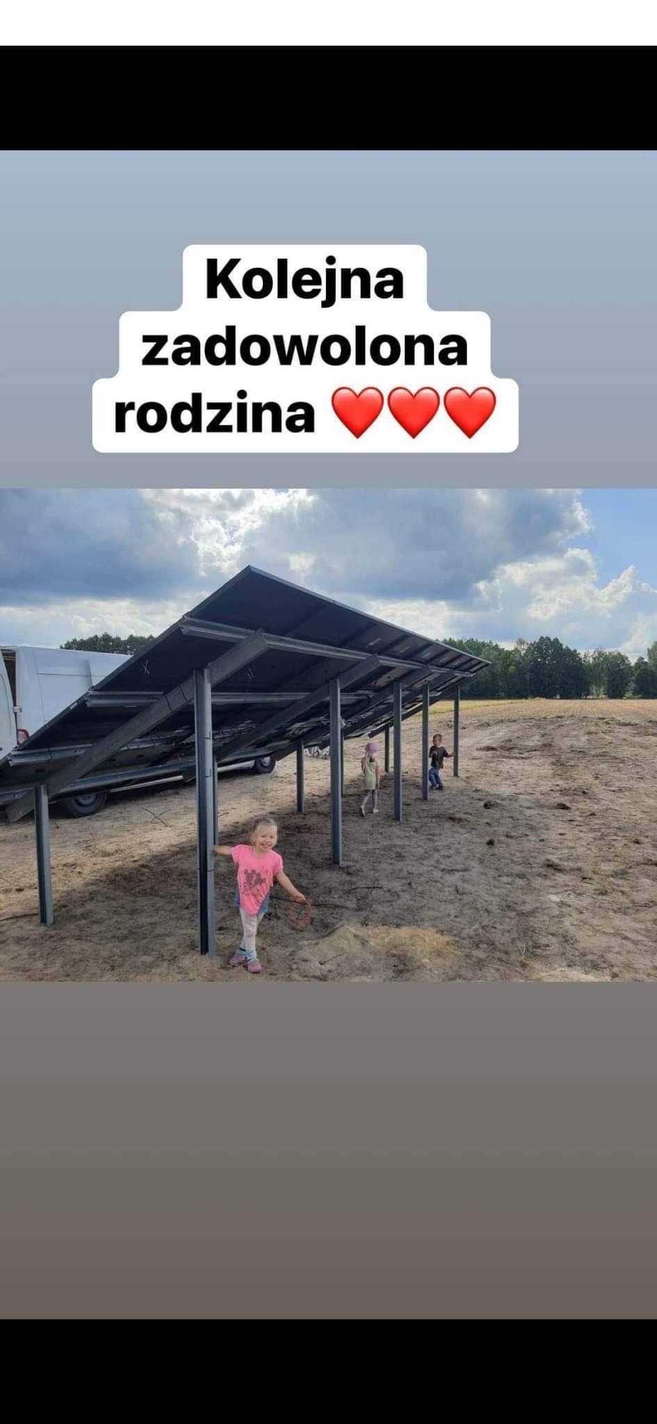 Uśmiechnięta dziewczynka stoi obok metalowych słupków konstrukcji pod panele słoneczne na tle dwóch bawiących się dzieci i częściowo widocznego białego samochodu dostawczego.