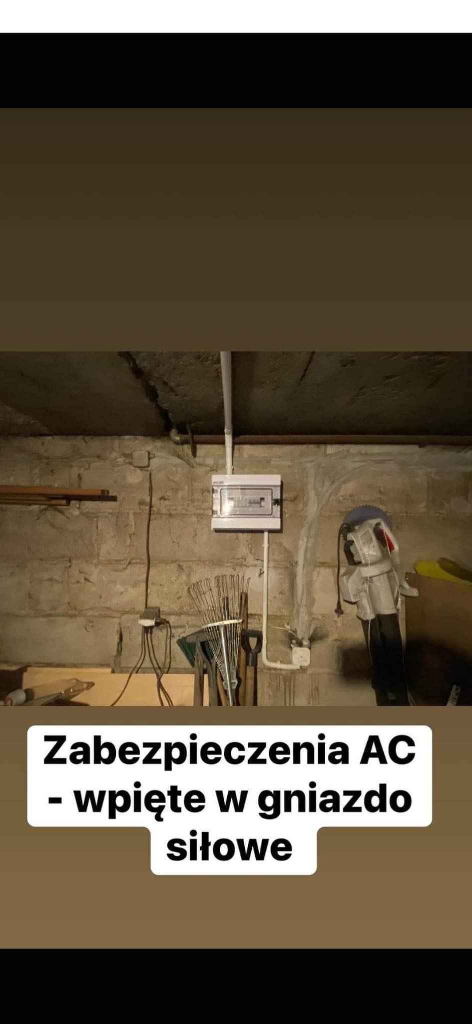 Skrzynka elektryczna z bezpiecznikami zamontowana na ceglanej ścianie piwnicy, widoczne rury instalacyjne, kabel zasilający i narzędzia ogrodowe w tle.