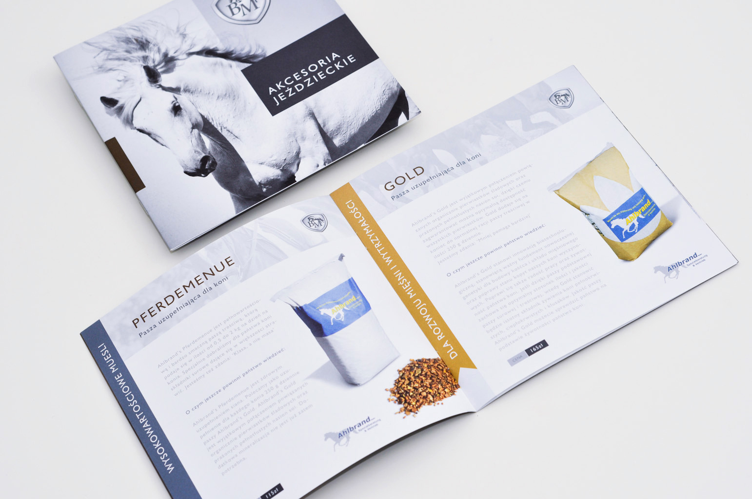 Rozkładówka firmowej broszury o akcesoriach jeździeckich i paszach dla koni, z widocznym zdjęciem białego konia i opakowaniami pasz Ahlbrand.