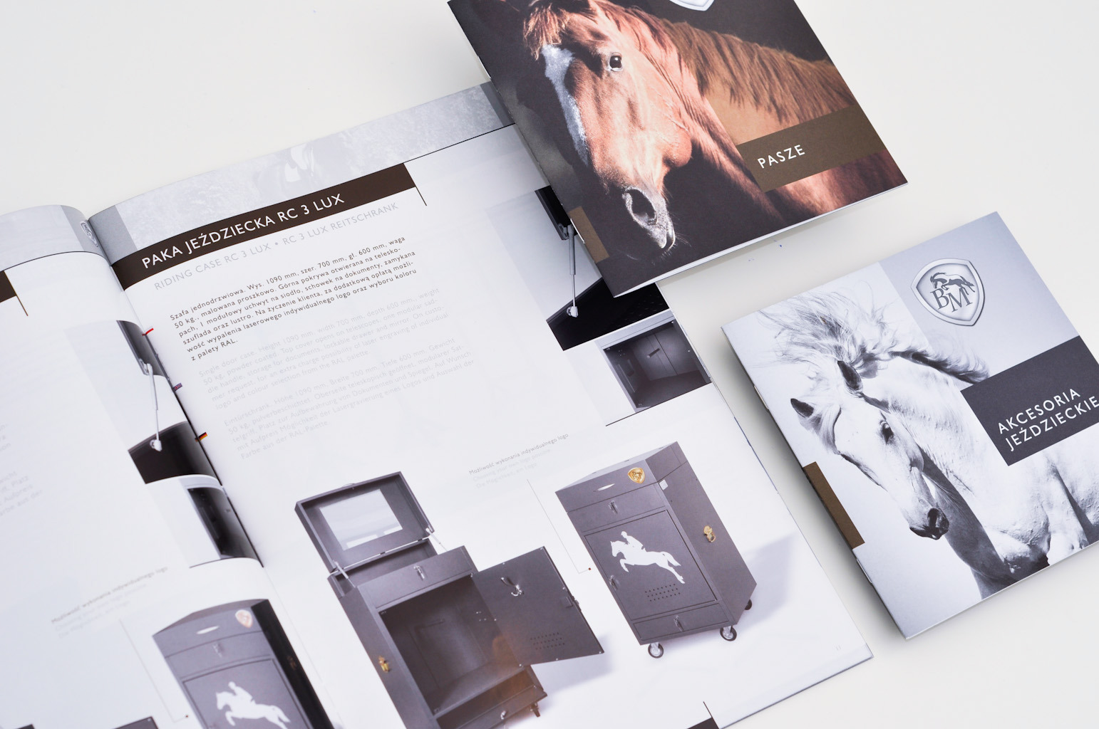 Katalog produktowy z otwartą stroną prezentującą szafę jeździecką na kółkach z otwartymi drzwiami i lustrem, obok leżą dwa zamknięte katalogi z motywami koni i akcesoriami jeździeckimi.