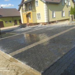 Fach-bruk - Świeżo ułożona, mokra kostka brukowa na podjeździe przed domem, pracownik zagęszcza powierzchnię wibratorem, w tle widoczny dom z balkonem i zielony budynek gospodarczy.