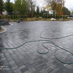 Fach-bruk - Świeżo ułożona, mokra kostka brukowa w odcieniach szarości, z widocznym wężem ogrodowym, fragmentem drewnianej konstrukcji i przygotowanym terenem pod dalsze prace ogrodowe w tle.