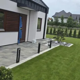 Nowoczesny ogród z kostką brukową, oświetleniem i trawnikiem. Widoczne elementy małej architektury i zadbana zieleń.