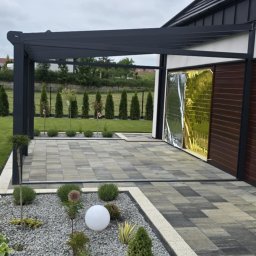 Fach-bruk - Nowoczesna pergola z kostki brukowej w szarościach, uzupełniona oświetleniem kulistym i rabatą z kamienia oraz roślin.  Widok na starannie utrzymany ogród.