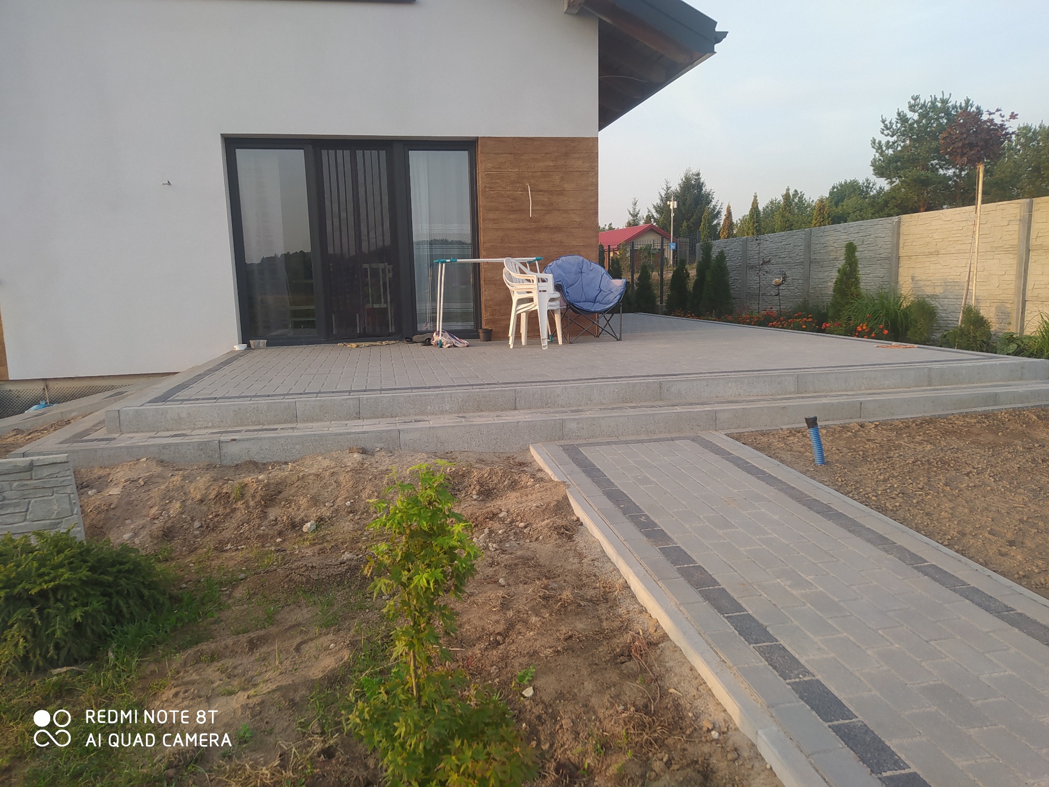 Nowo ułożony chodnik z kostki brukowej prowadzący do tarasu przed nowoczesnym domem z białymi ścianami i drewnianą okładziną, na tarasie ustawione krzesła i suszarka na pranie.