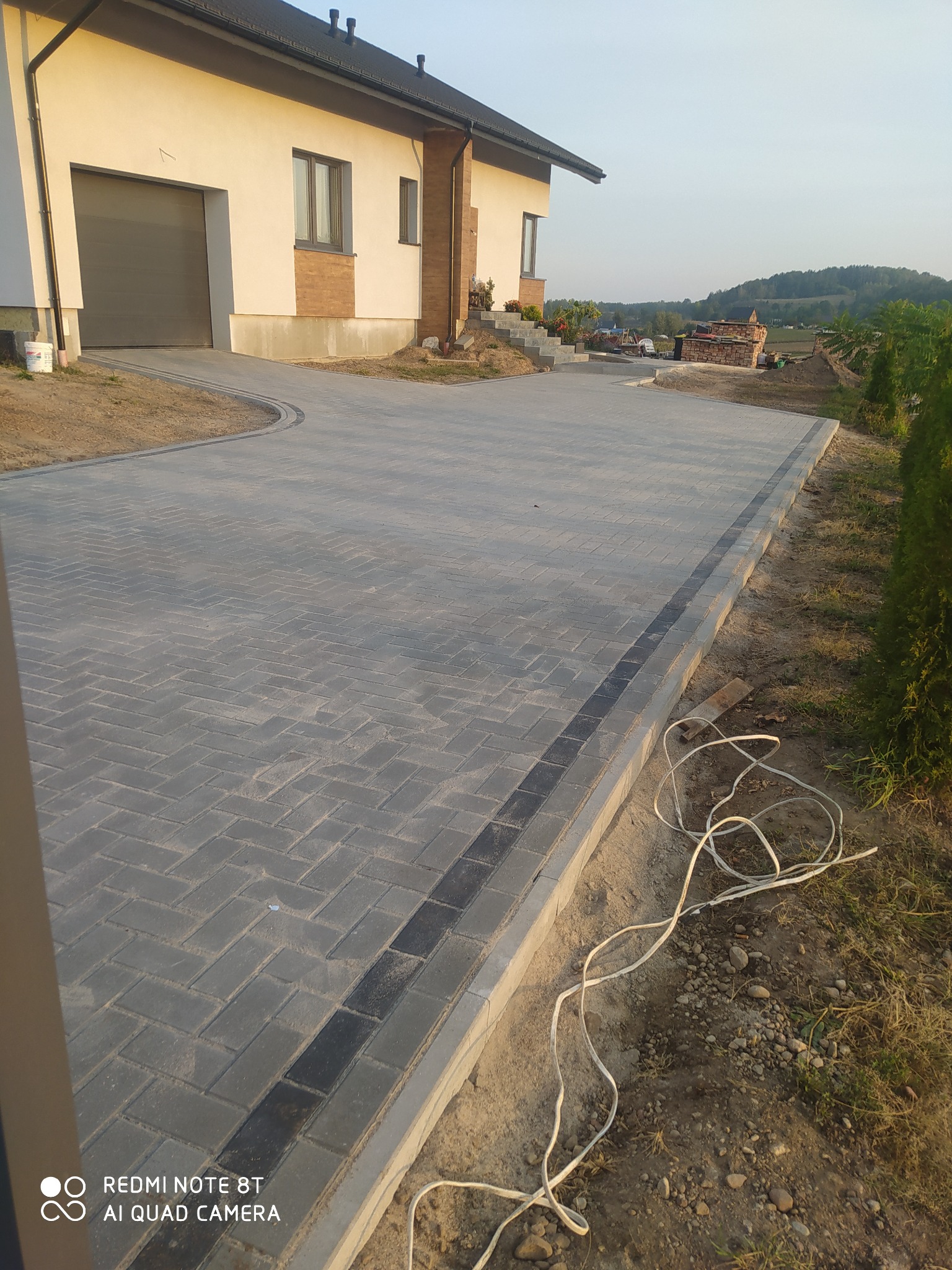 Szeroki podjazd z szarej kostki brukowej ułożonej w jodełkę, z ciemniejszym obramowaniem, prowadzący do nowoczesnego domu z garażem; w tle widok na pagórkowaty krajobraz.