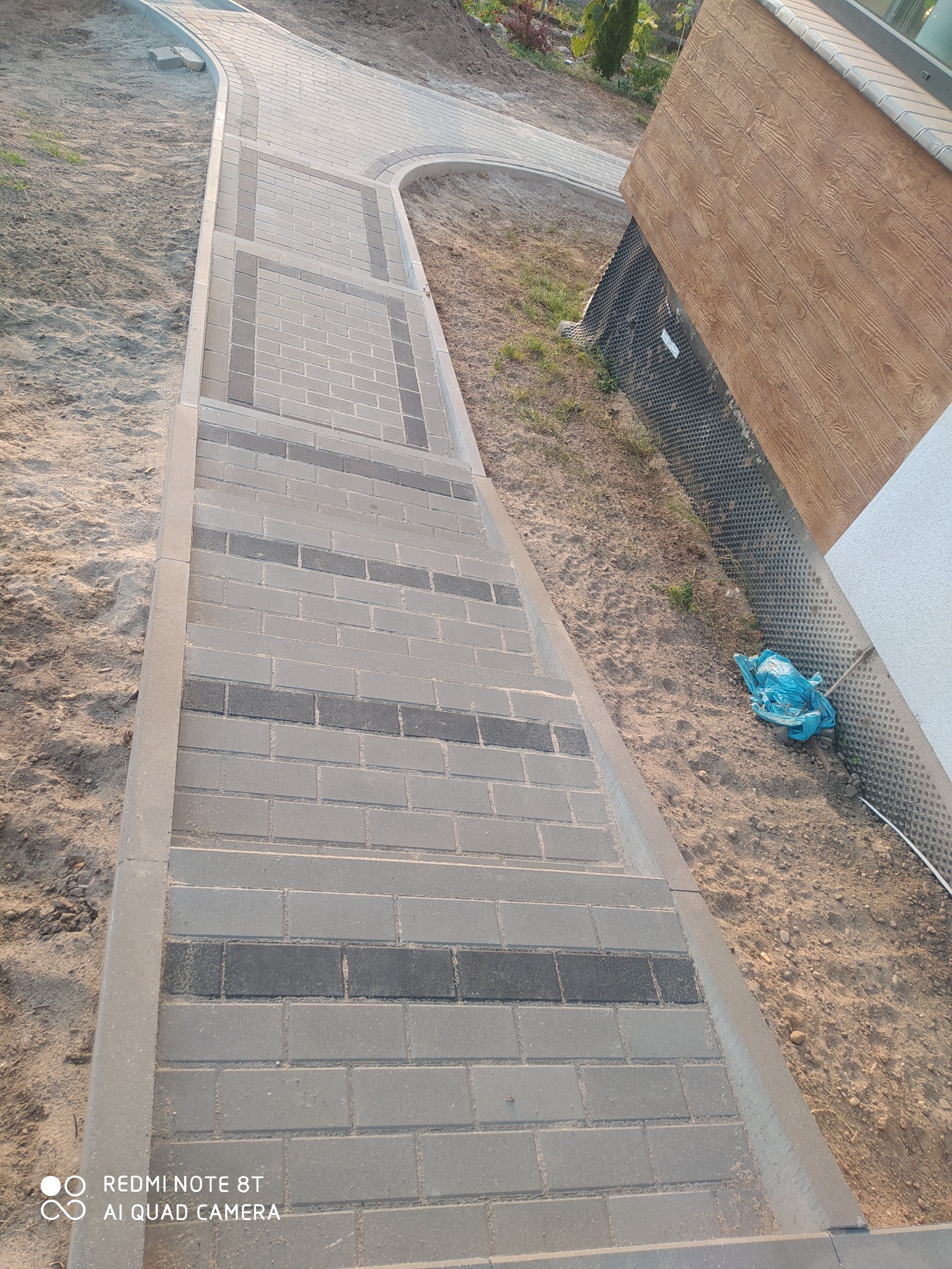 Świeżo ułożona ścieżka z kostki brukowej w odcieniach szarości, z ciemniejszymi akcentami, prowadząca wzdłuż budynku z drewnianą elewacją i zabezpieczeniem fundamentów, widoczny fragment trawnika...