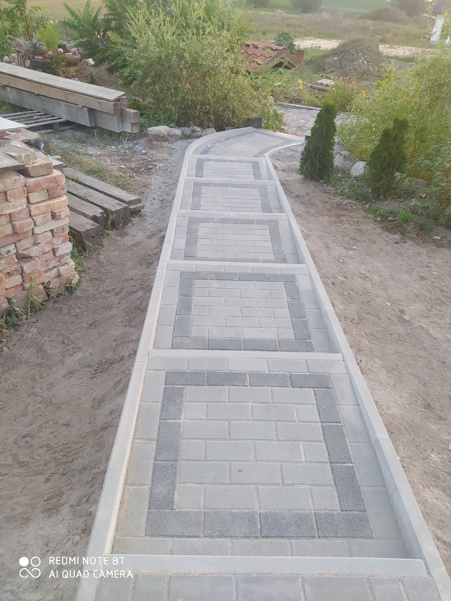 Ułożona ścieżka z kostki brukowej w odcieniach szarości prowadząca przez ogród, z widocznymi przygotowanymi cegłami i deskami po lewej stronie, sugerującymi trwające prace.