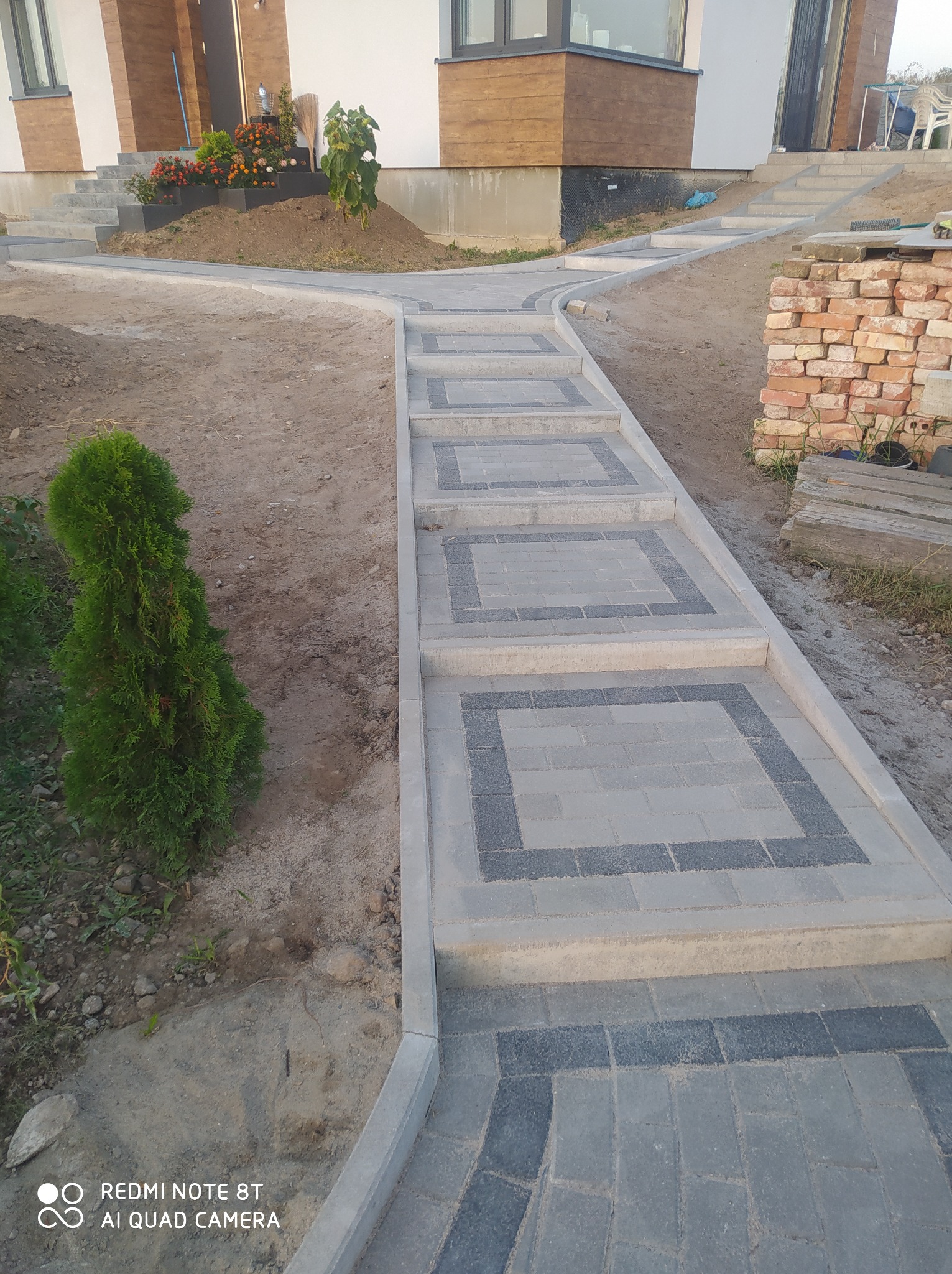 Nowo ułożona ścieżka z kostki brukowej w dwóch odcieniach szarości, prowadząca do domu z rabatami kwiatowymi, widoczna mała tuja obok ścieżki.