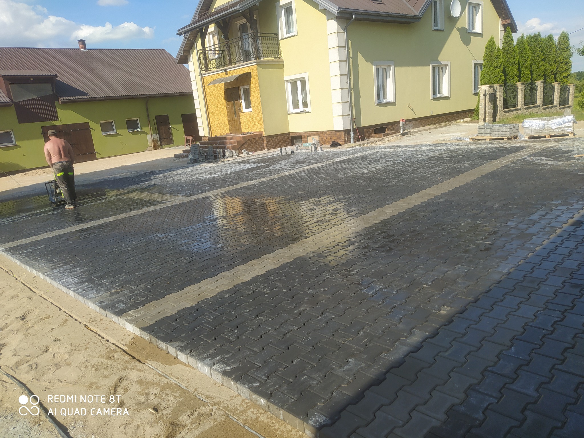 Świeżo ułożona, mokra kostka brukowa na podjeździe przed domem, pracownik zagęszcza powierzchnię wibratorem, w tle widoczny dom z balkonem i zielony budynek gospodarczy.