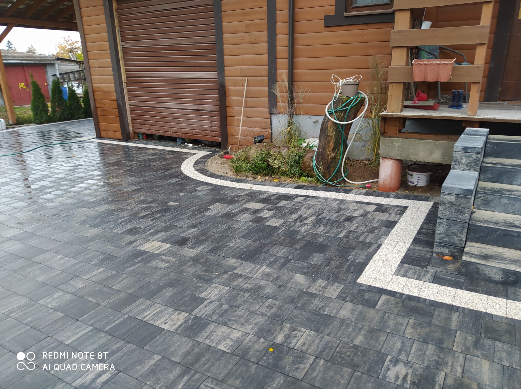 Ułożona szara kostka brukowa z białym obramowaniem przy drewnianym budynku z roletą, widoczny pień drzewa z wężem ogrodowym i schody z kostki brukowej.