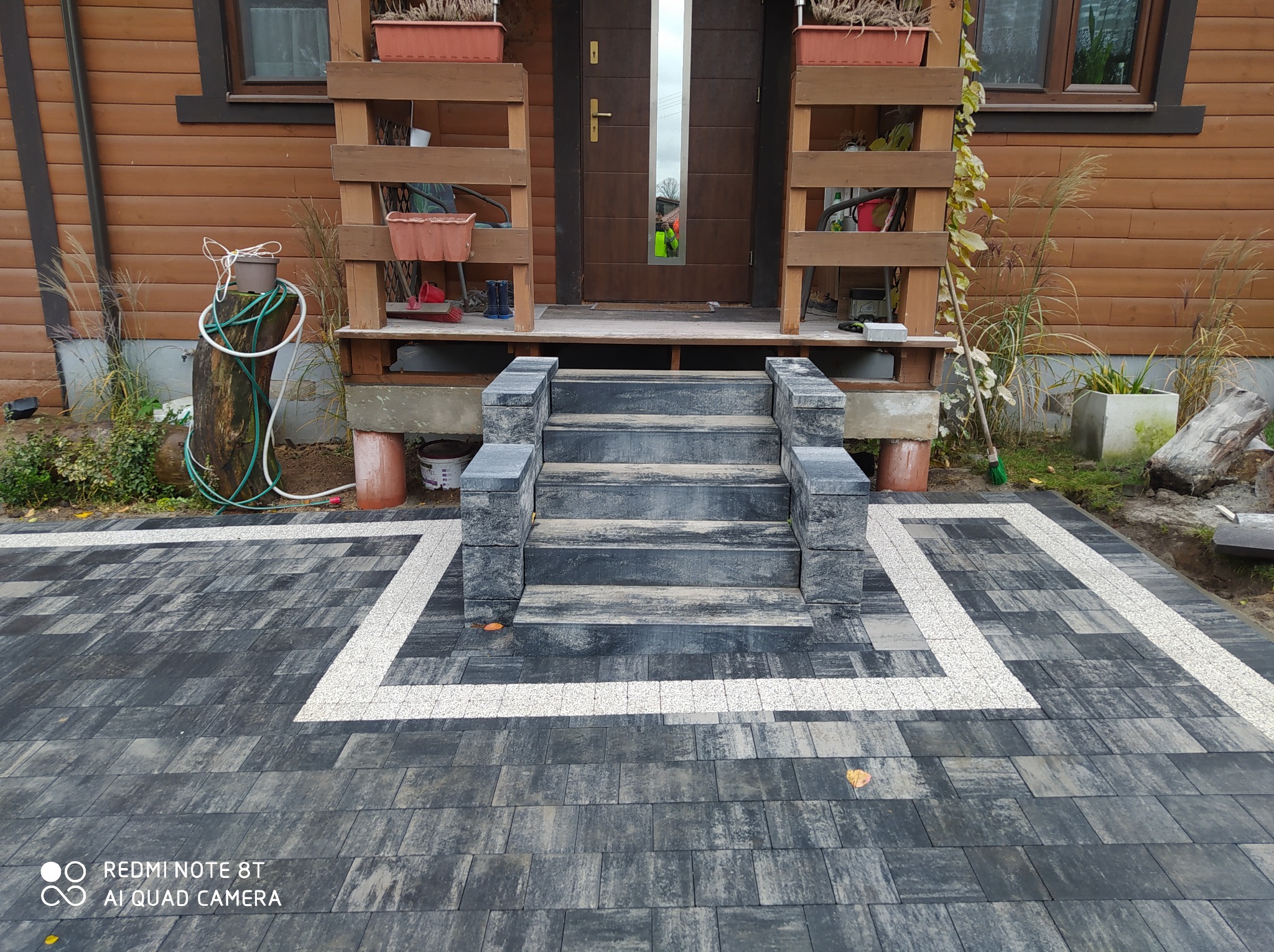 Nowo ułożona, szara kostka brukowa z białym obramowaniem prowadząca do drewnianego domu z niewielkimi schodami wejściowymi. Widoczne donice z roślinami na werandzie.
