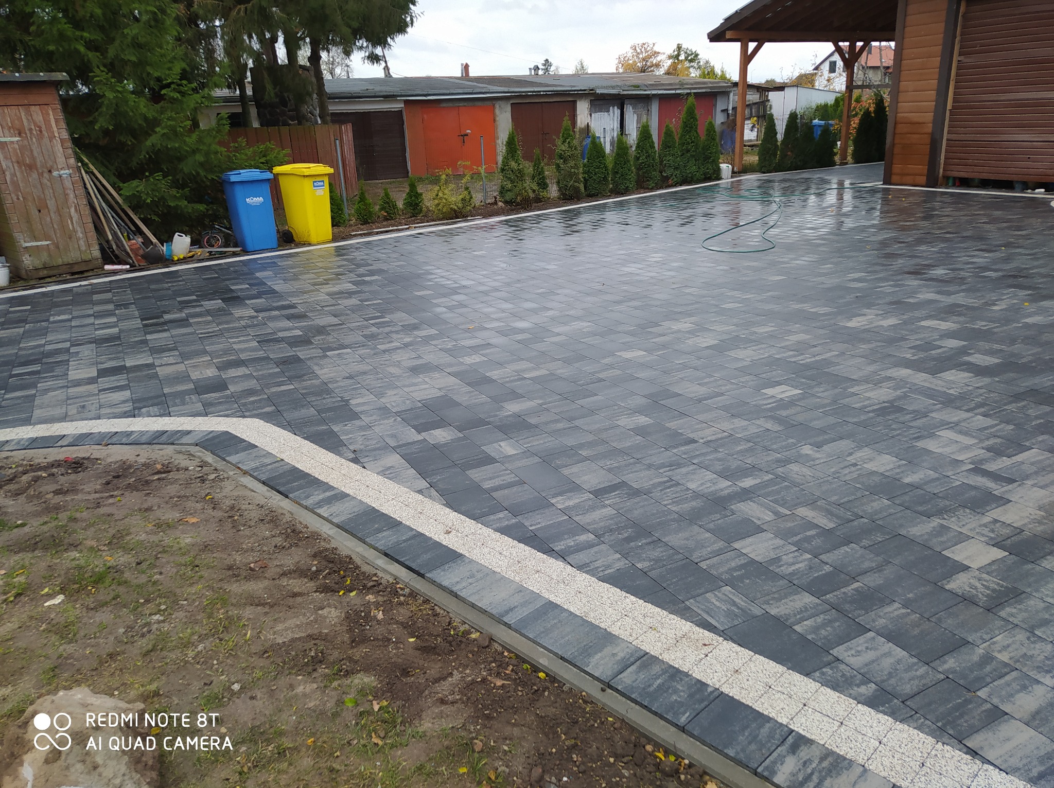 Ułożona kostka brukowa w kolorze szarości, z obrzeżem z jasnego kruszywa, widok na posesję z garażami i zielenią w tle, lekko pochmurne niebo.
