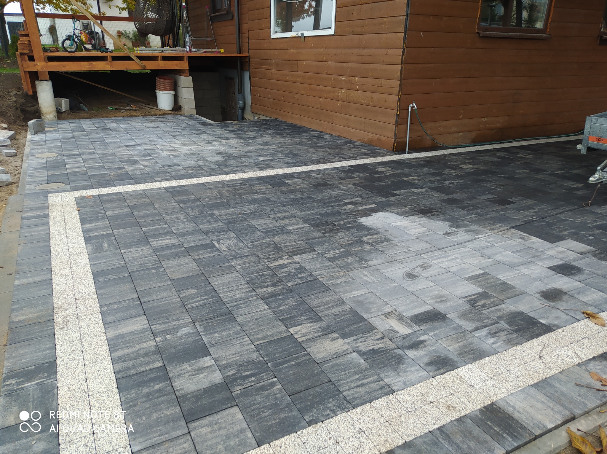 Ułożona, szara kostka brukowa z jasnymi paskami dekoracyjnymi, przylegająca do drewnianego budynku z widocznymi elementami ogrodu i podestu.