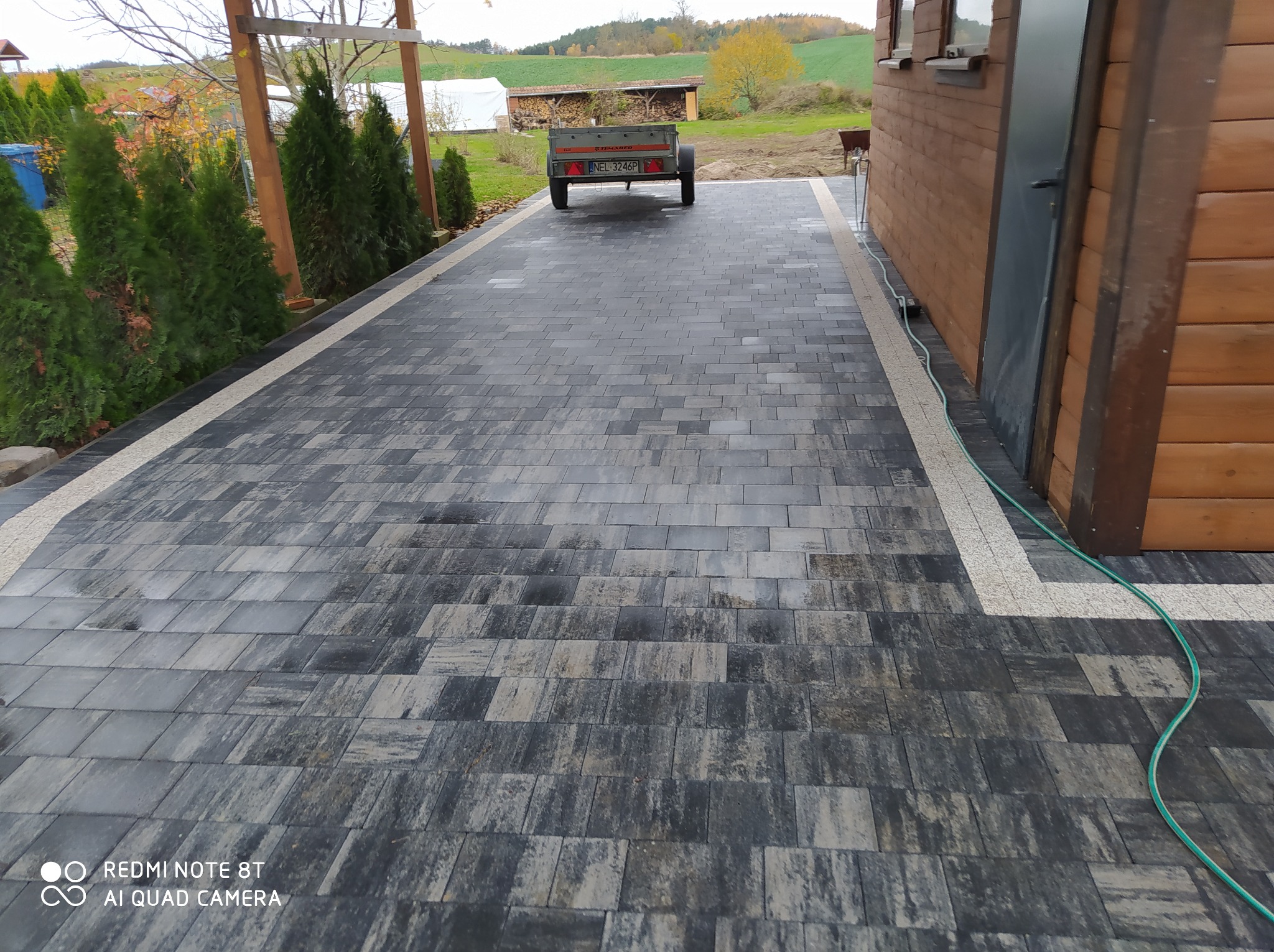 Szeroki podjazd z szarej kostki brukowej o nieregularnym wzorze, obramowany jasnymi pasami, prowadzący do drewnianego domku z przyczepką w tle, widok z perspektywy lekko z góry.