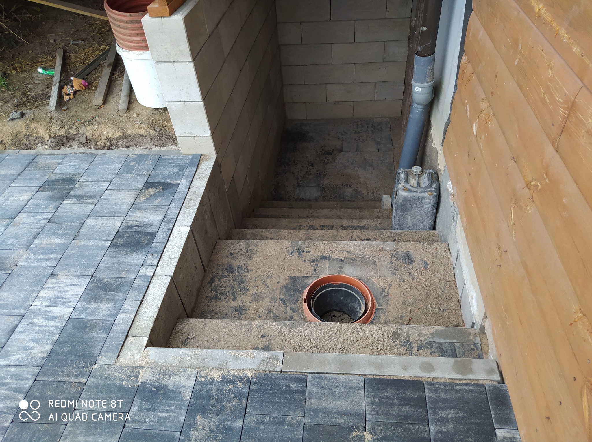 Schody z kostki brukowej w trakcie budowy, widoczne przygotowane podłoże z piasku, elementy odwodnienia i fragment drewnianej elewacji budynku.