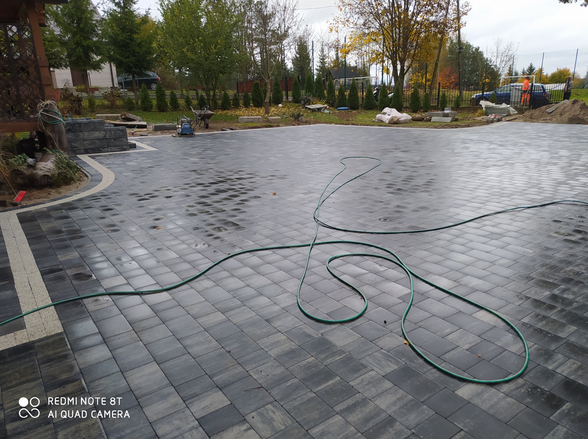 Świeżo ułożona, mokra kostka brukowa w odcieniach szarości, z widocznym wężem ogrodowym, fragmentem drewnianej konstrukcji i przygotowanym terenem pod dalsze prace ogrodowe w tle.