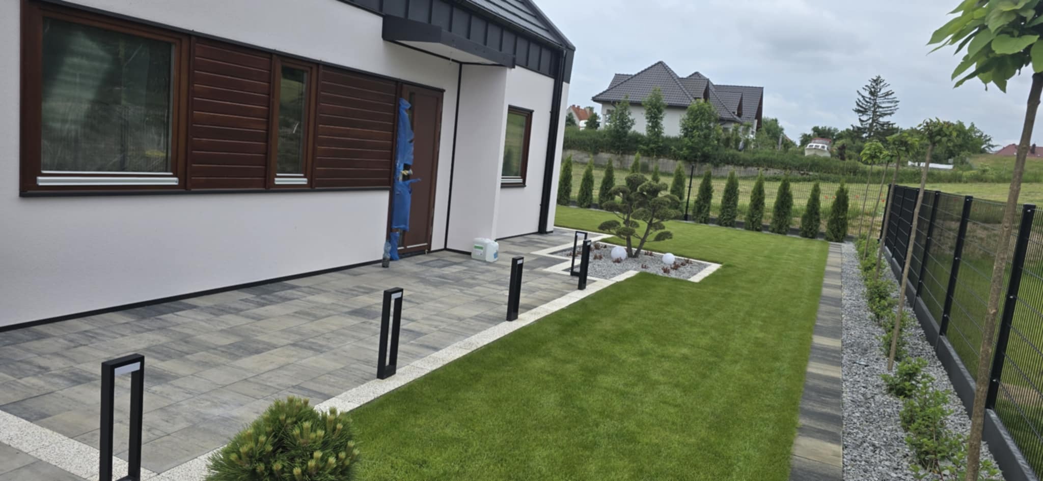 Nowoczesny ogród z kostką brukową, oświetleniem i trawnikiem. Widoczne elementy małej architektury i zadbana zieleń.