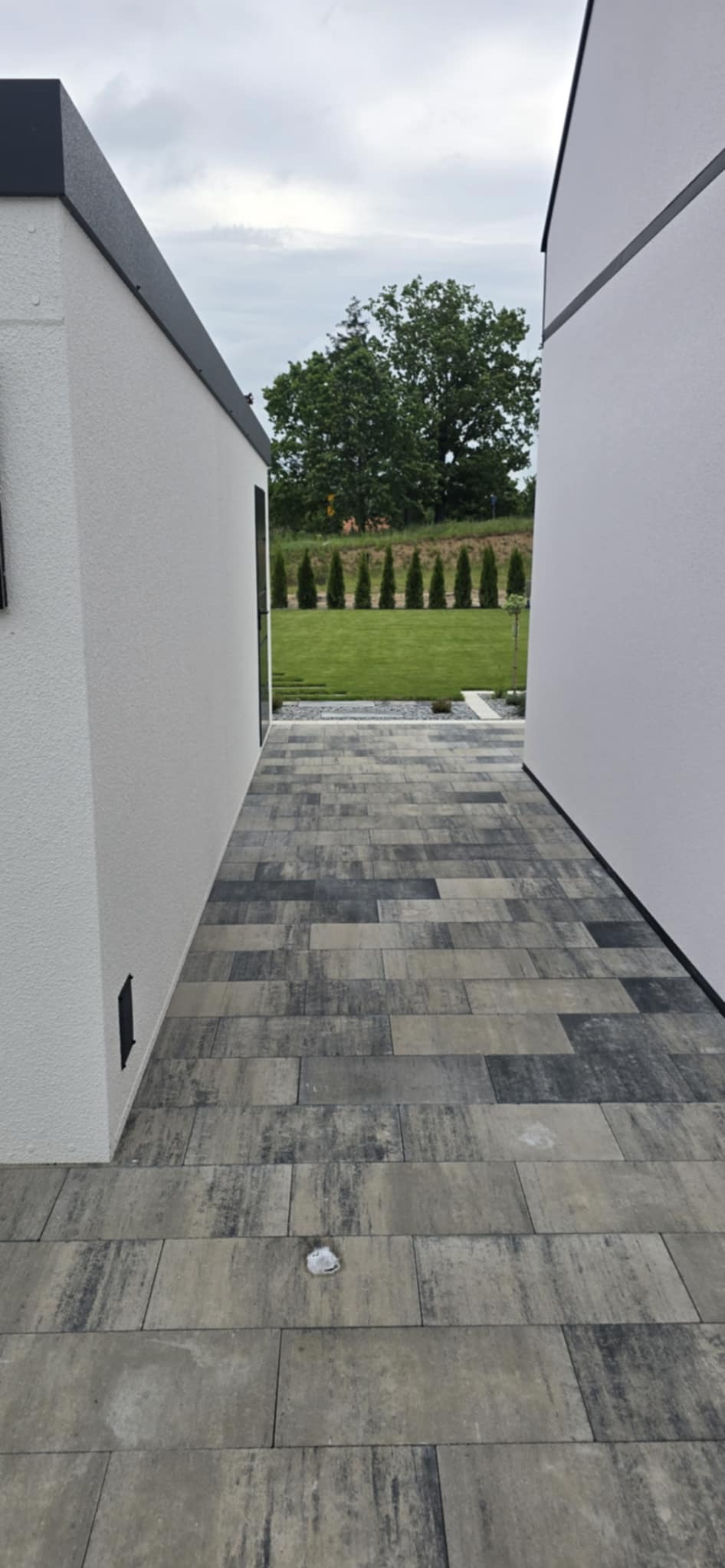 Wyłożona kostką brukową ścieżka między budynkami, prowadząca do zadbanego ogrodu z równo posadzonymi drzewami. Kostka w odcieniach szarości i czerni.
